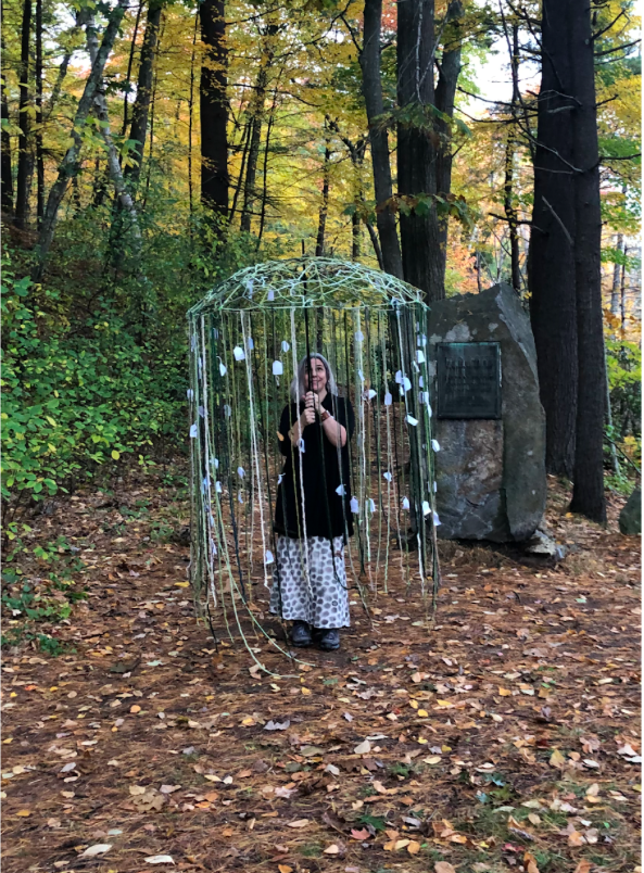 karen Krolak is walking in the woods under and enormous sculptural umbrella with long crocheted tendrils hanging from it. Pieces of paper are tied to the tendrils.