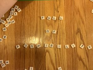 Still from the film Pane #4302.1.  Bananagram letters are scattered on a wooden floor. Some of them spell out the words, Full Of Possibility
